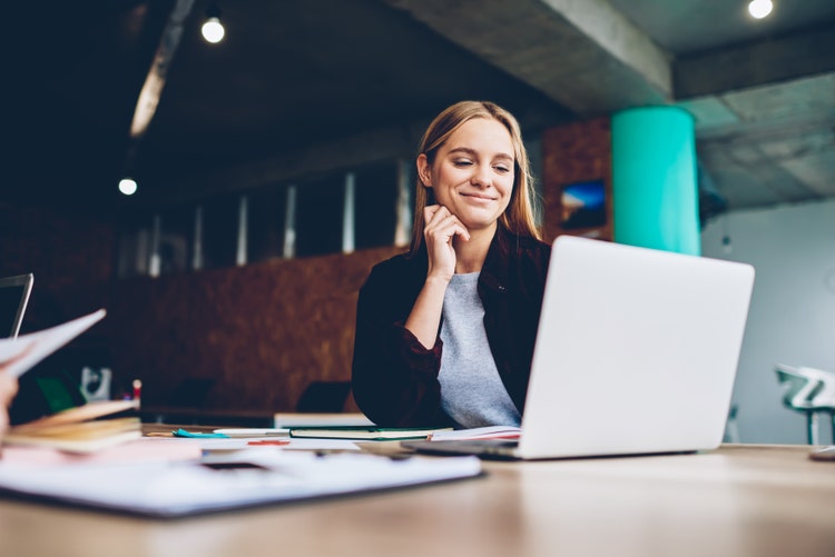 A smiling woman sits at a cluttered desk in front of a laptop, resting her chin on one hand.
