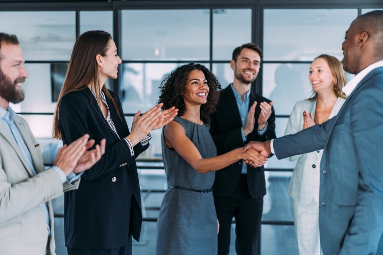 Two businesspeople shaking hands, surrounded by a group of other suited professionals in a corporate setting