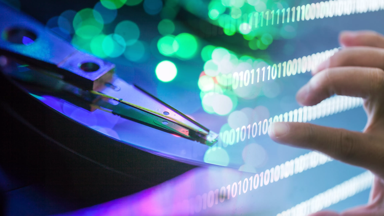 A close-up of a human finger delicately touching a screen displaying binary code, with a faded hard disk drive gently blurred in the background.