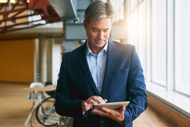 An executive in a business suit stands indoors near large windows, holding and using a tablet device.