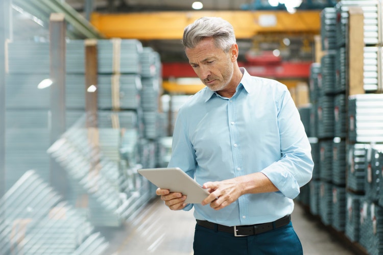 An older executive male in a button-up shirt and dark pants looks at a tablet while standing a warehouse.