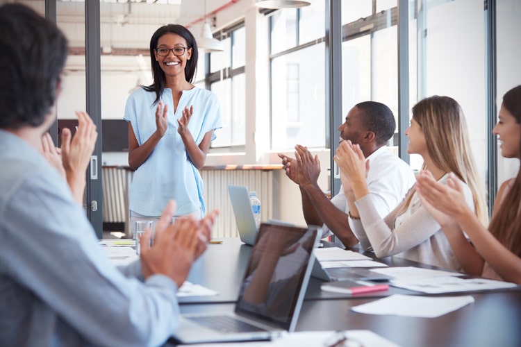 Young black woman stands clapping with colleagues in a meeting.