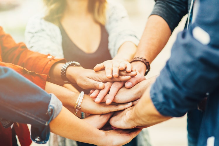 A diverse group of people stand in a circle with their hands stacked on top of each other in the center, symbolizing unity and teamwork.