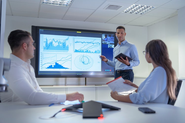 Person holding tablet presents data charts on a screen to two seated colleagues in conference room.