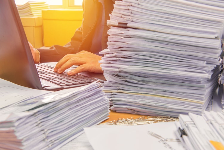 A person typing on a laptop surrounded by stacks of documents on the desk