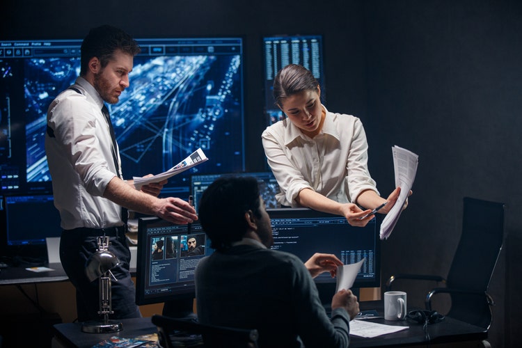 Three people are in a dimly lit office with multiple computer screens displaying data and images, having a conversation.