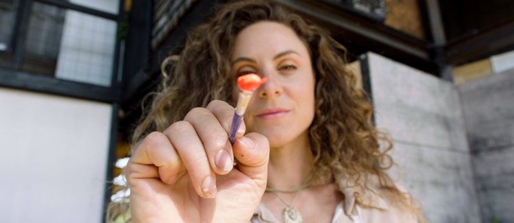 Woman with curly hair holding up a dollop of orange paint on the end of a paintbrush toward the camera.
