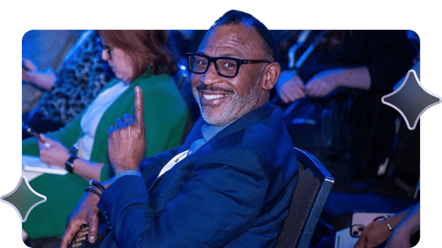 Man in a blue suit smiling and pointing upward while seated at a conference. The mood is positive, with people in the background.