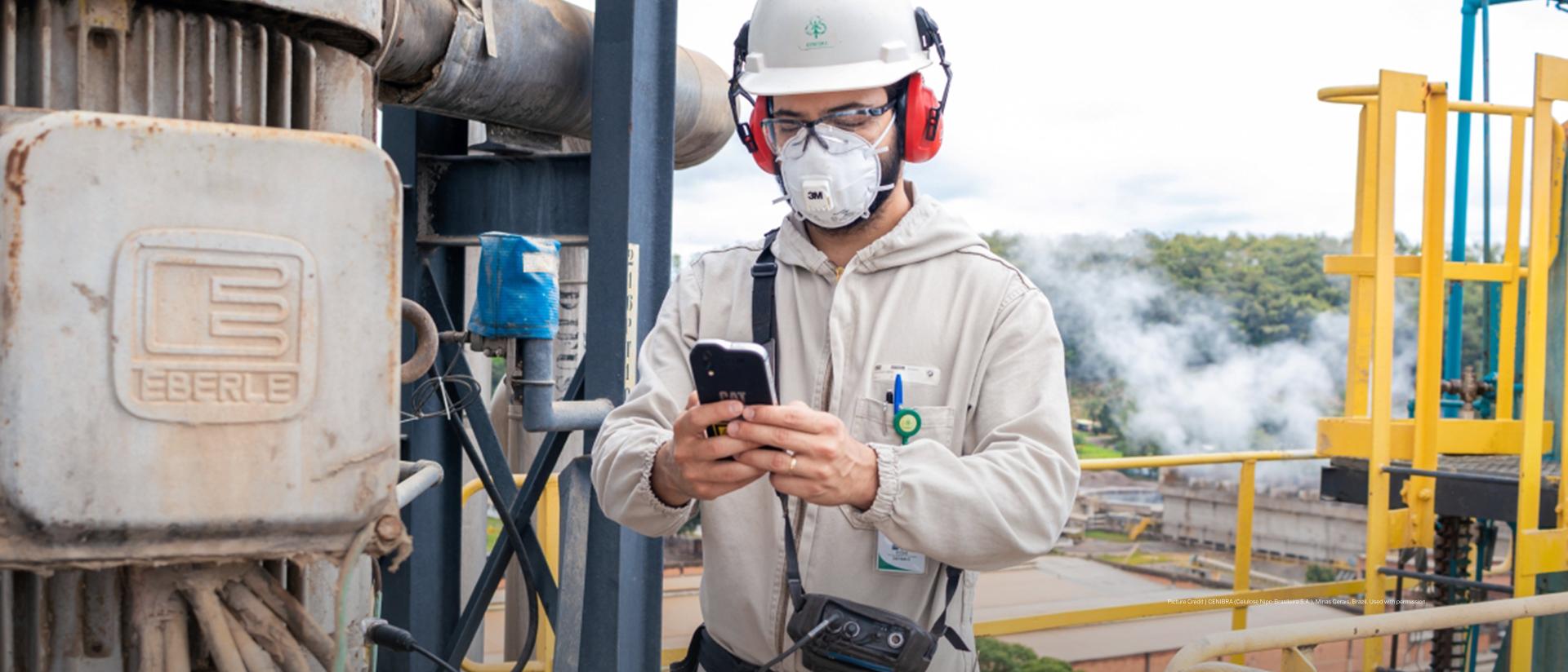 CENIBRA worker with protective gear on site of a pulp plant holding a mobile device