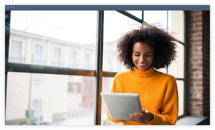 A smiling woman standing in front of a window and looking at a tablet.