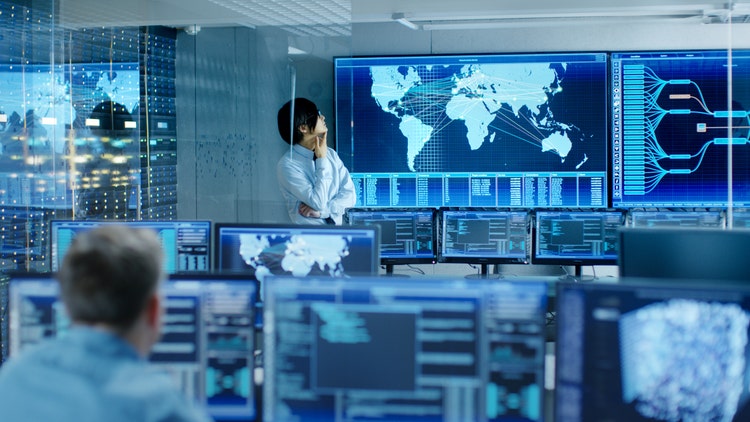A room with monitors displaying data and maps. A businessperson looks at the world map on a large screen. Another person works on screens