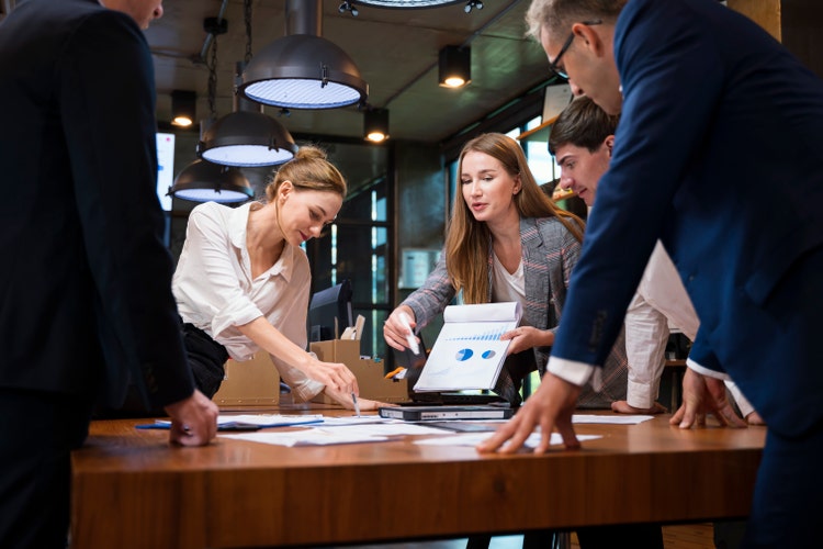 A team of professionals lean around a boardroom table to review paper printouts of complex business data