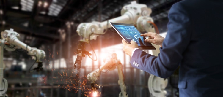 An engineer overseeing automated robot arms in a smart factory using real-time monitoring software on a tablet to control and check welding robotics and digital manufacturing processes