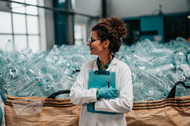 A woman employee, holding a clipboard, stands in front of a large, overflowing basket filled with empty, used plastic water bottles at a recycling plant.