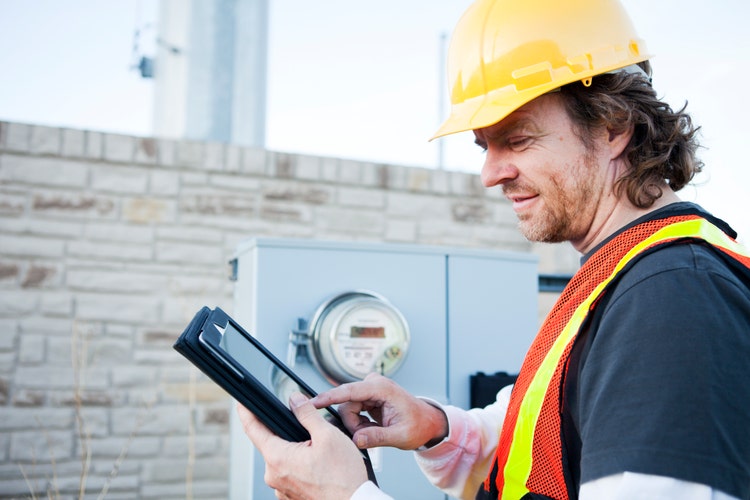 A power line technician wearing a yellow hard hat and a safety vest and using a tablet while standing in front of a meter box