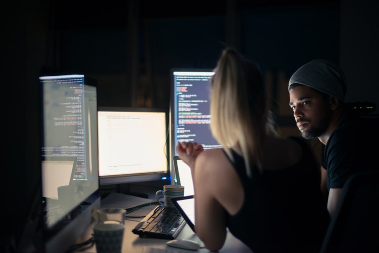 Two coders in a darkly lit room looking at various computer monitors with code